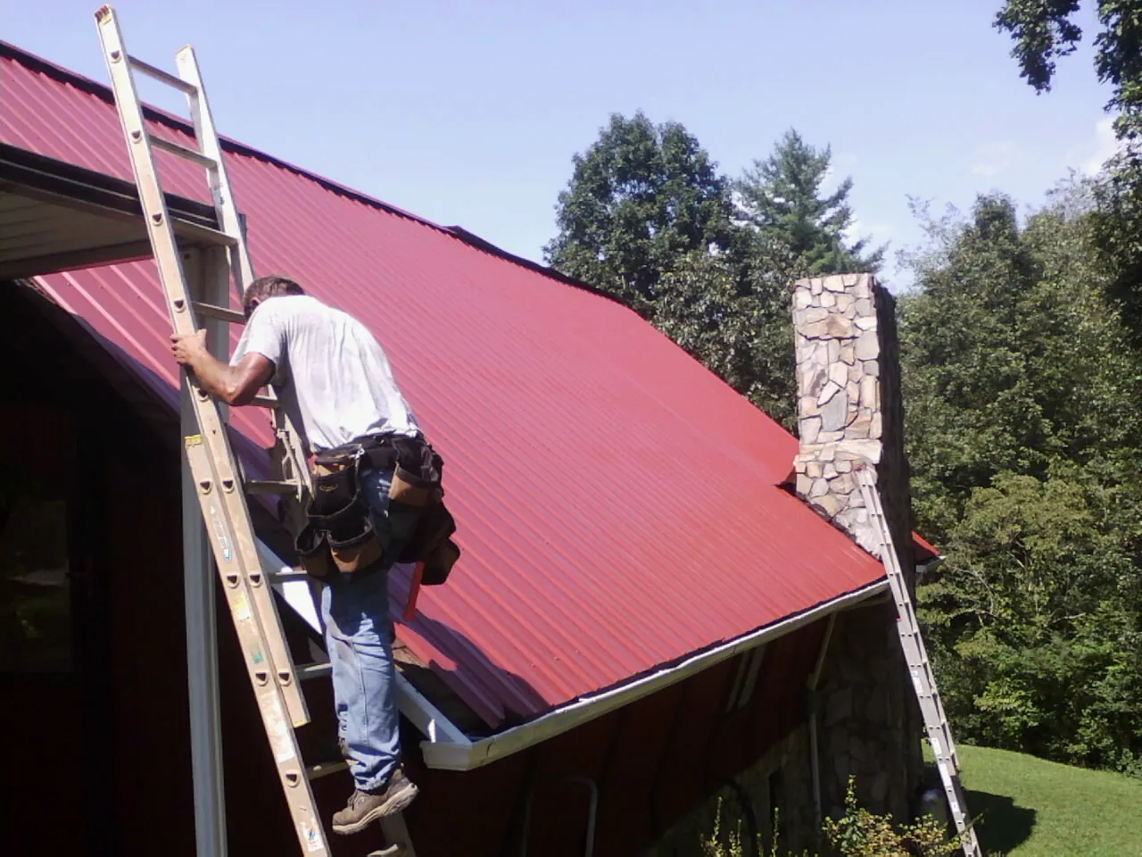 Roofer on a ladder beside a red metal roof and stone chimney — Soffit & Fascia Repair by Roofing Brothers in Kingston Mines